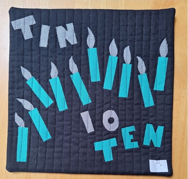 A square quilt with a black background and a gray border. The quilt features the word "TEN" written in gray letters, surrounded by a row of teal candles. The background is quilted with a linear pattern.