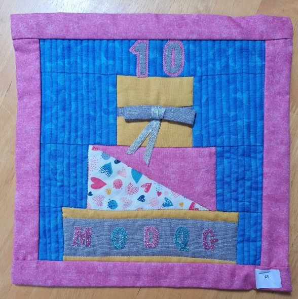 A square quilt with a pink background and a pink border. The quilt features a birthday cake with the number "10" on top, a yellow box with a ribbon, and the word "MODQG" embroidered on the bottom. The background is quilted with a linear pattern.