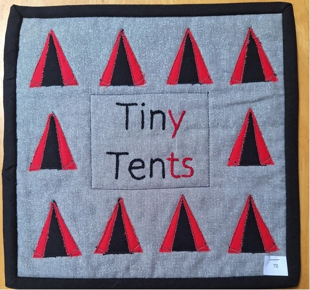 A square quilt with a gray background and a black border. The quilt features a pattern of nine small, triangular tents made from black and red fabric. The word "Tiny Tents" is embroidered in the centre.