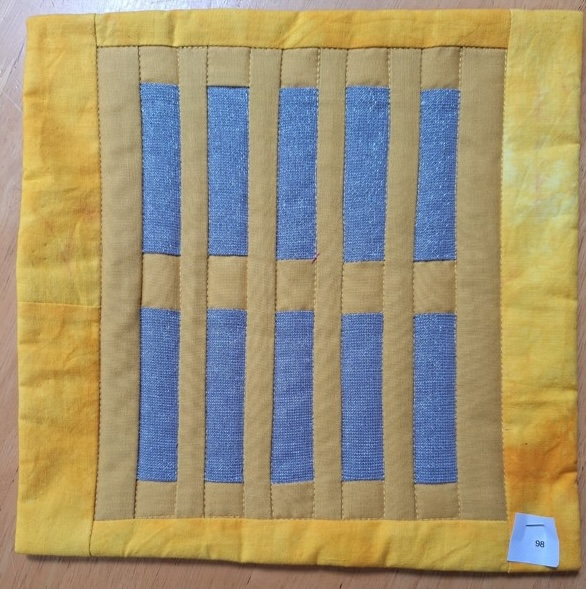 A square quilt with a yellow background and a yellow border. The quilt features a pattern of vertical gray lines, creating a sense of depth and texture.