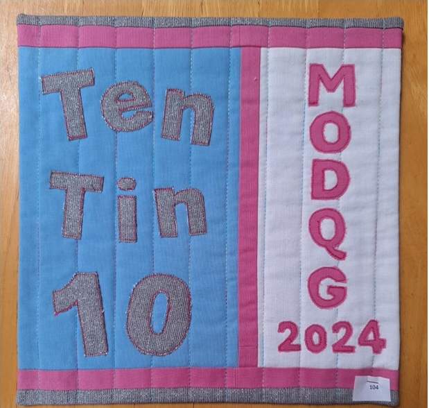 A square quilt with a blue background and a pink border. The quilt features the text "Ten Tin 10" and "MODQG 2024" in black and white letters. The background is quilted with a linear pattern.