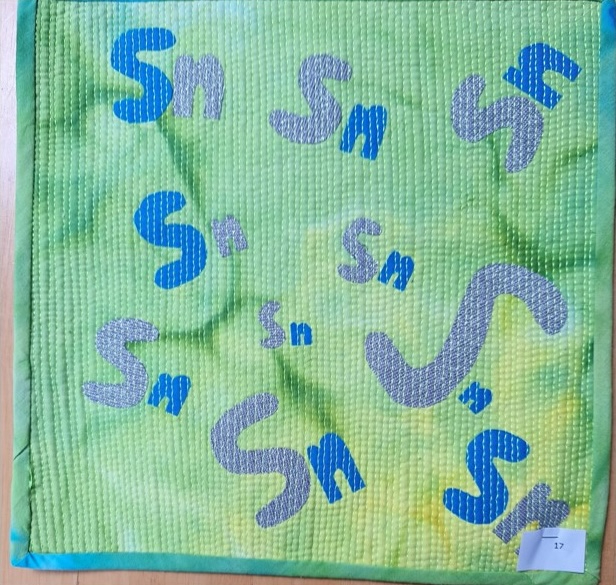 A square quilt with a green background and a blue border. The quilt features the letter "S" repeated multiple times in various sizes and colours, including gray and blue. The background is quilted with matchstick quilting