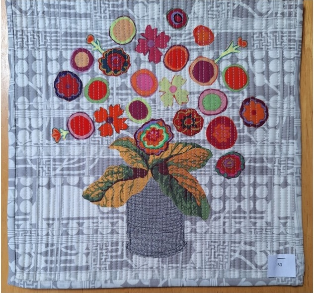 A square quilt with a gray background and a gray border. The quilt features a vase filled with colourful flowers, each made from different fabrics with various patterns. The background is quilted with a linear pattern.