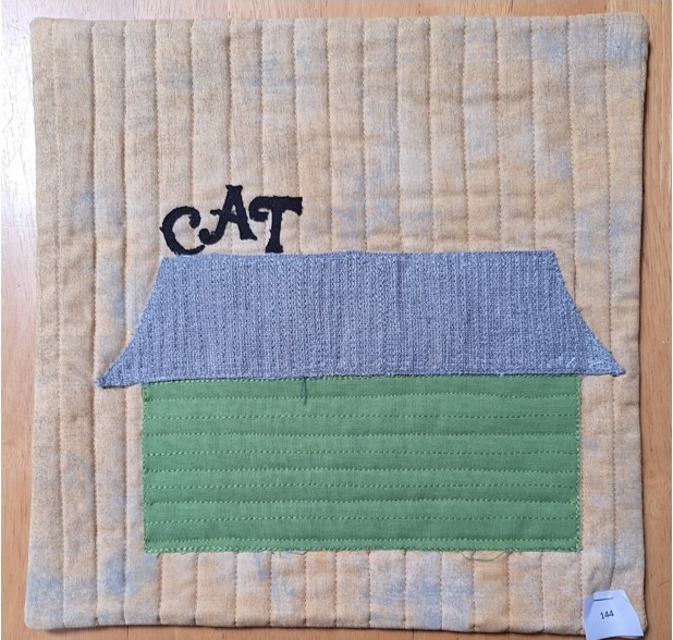 A square quilt with a yellow background. The quilt features a green house shape with a blue roof, and the word "CAT" embroidered on the roof. The background is quilted with vertical lines.