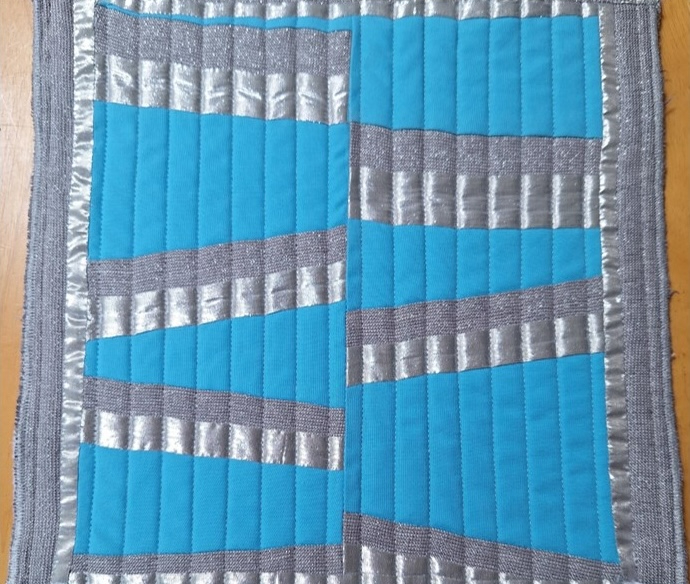 A square quilt with a blue background. The quilt features a pattern of horizontal, silver-coloured rectangles on the blue background. The background is quilted with a linear pattern.