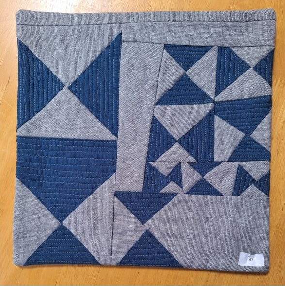 A square quilt with a gray background and a gray border. The quilt features a geometric design with triangles in shades of blue and gray. The triangles are arranged in a repeating pattern, creating a sense of rhythm and symmetry.