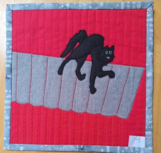 A square quilt with a red background and a gray border. The quilt features a black cat silhouette walking along a gray, textured roof. The background is quilted with vertical lines.