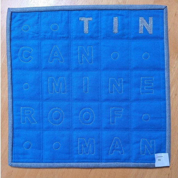 A square quilt with a blue background and a gray border. The quilt features a word search puzzle with the words "TIN CAN MINE ROOF MAN" spelled out in gray letters. The background is quilted with a linear pattern.