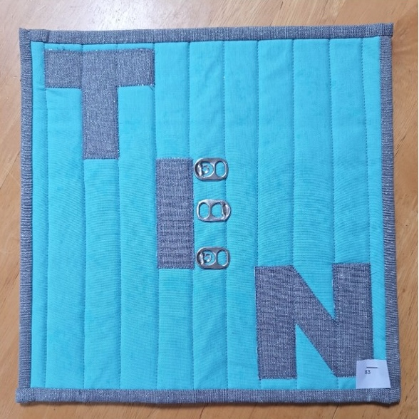 A square quilt with a turquoise background and a gray border. The word "TIN" is spelled out in gray letters, with the letter "I" also creating an E by a row of four tin can tabs. The background is quilted with vertical lines.