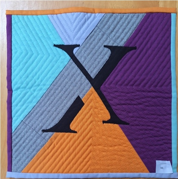 A square quilt with a colourful background and a gray border. The quilt features a large, black letter "X" in the centre. The background is divided into four sections in turquoise, gray, orange, and purple. Each section is quilted with a chevron pattern.