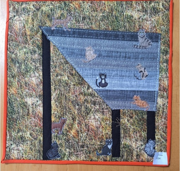 A square quilt with an orange border. The quilt features a table with several cats sitting on it and around it. The cats are made of different fabrics with various patterns. The background is a textured, brown fabric. The quilt is quilted with a linear pattern.