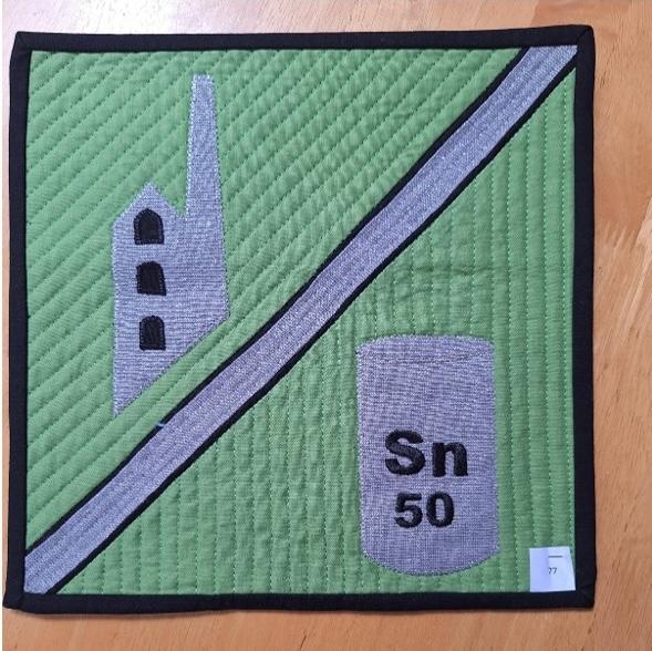 A square quilt with a green background and a black border. The quilt features a gray factory building on the left side and a gray tin can with the symbol "Sn" and the number "50" on the right side. A gray line separates the two objects. The background is quilted with a linear pattern.