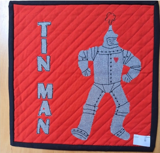 A square quilt with a red background and a black border. The quilt features the Tin Man from The Wizard of Oz, standing on a red background. The words "TIN MAN" are embroidered vertically on the left side. The background is quilted with a diagonal line pattern.