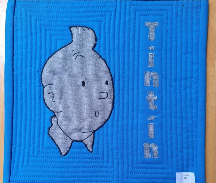 A square quilt with a blue background. The quilt features an embroidered image of Tintin, the famous comic book character, with his signature quiff and inquisitive expression. The word "Tintin" is embroidered vertically beside him. The background is quilted with a linear pattern.