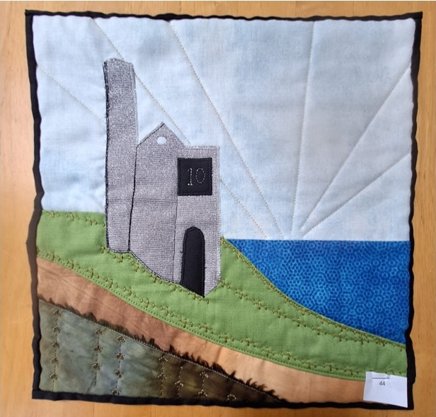 A square quilt with a blue background and a brown border. The quilt features a gray building with a tower on a green hill overlooking a blue body of water. The sky is depicted with white and blue fabric.