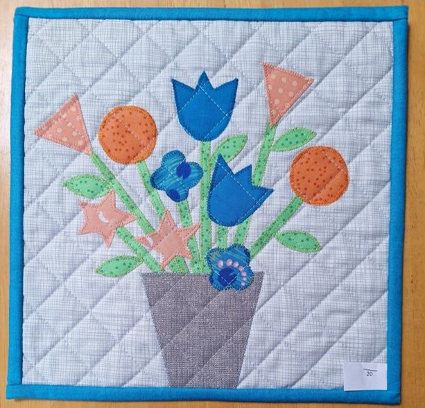 A square quilt with a white background and a blue border. The quilt features a colourful bouquet of flowers in a gray pot. The flowers include tulips, daisies, and other abstract shapes, all made from different fabrics with various patterns. The background is quilted with a hatched pattern.