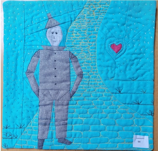 A square quilt with a blue background and a yellow border. The quilt features the Tin Man from The Wizard of Oz, standing on a yellow brick road. A red heart is visible in the background. The background is quilted with a linear pattern.