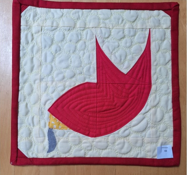 A square quilt with a red background and a red border. The quilt features a red fish shape with a yellow and blue tail. The background is filled with small, white circles