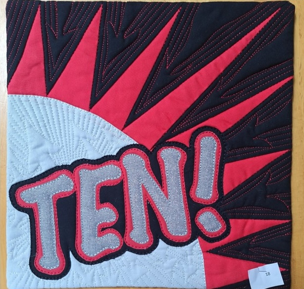 A square quilt with a red background and a black border. The word "TEN!" is written in gray letters with a comic book style speech bubble effect. The background is quilted with a radiating line pattern.