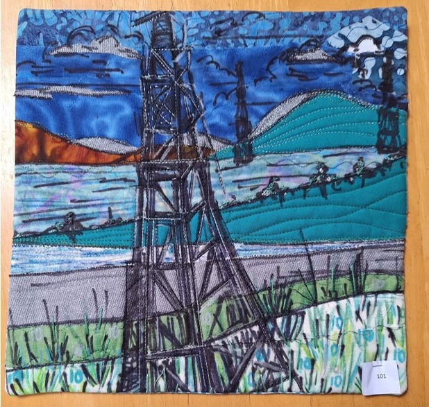A square quilt depicting a coastal landscape. The quilt features a large oil rig in the foreground, with a mountainous landscape and a cloudy sky in the background. The scene is made up of various fabric pieces with different textures and patterns, creating a mixed-media effect.