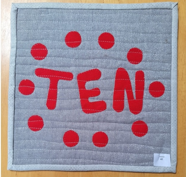 A square quilt with a gray background and a red border. The quilt features the word "TEN" spelled out in red letters, surrounded by a circle of red dots.