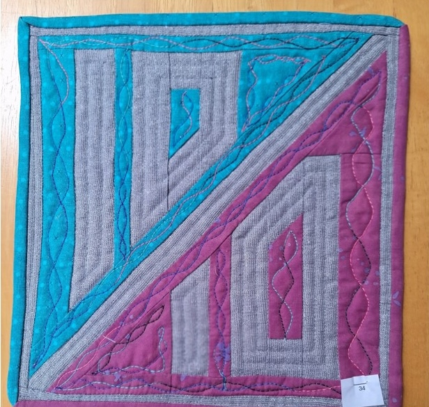 A square quilt with a gray background and a colourful border. The quilt is divided diagonally into two sections: one turquoise and the other pink. Both sections feature a maze-like pattern in gray. The background is quilted with intertwining wavy lines.