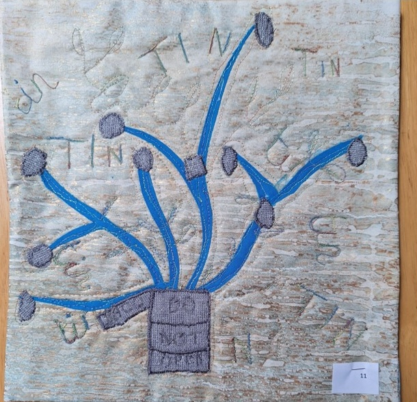 A square quilt with a light blue background and a gray border. The quilt features a tree-like structure with blue branches and gray leaves. The word "TIN" is embroidered on the branches and leaves.