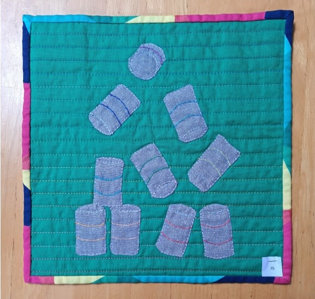 A square quilt with a green background and a colourful border. The quilt features several grey, cylindrical shapes representing cans, arranged in a playful, scattered pattern. The background is quilted with a linear pattern.