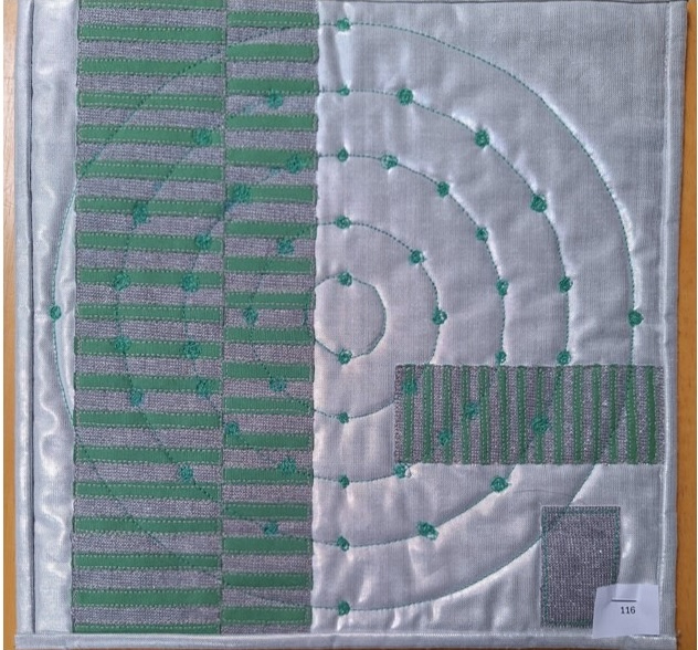 A square quilt with a gray background and a gray border. The quilt features a circular design with concentric circles in the centre. On the left side, there is a vertical section with horizontal lines and small, circular patterns.