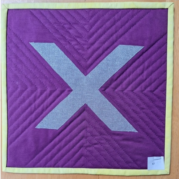 A square quilt with a purple background and a yellow border. The quilt features a large, gray letter "X" in the centre. The background is quilted with a chevron pattern.