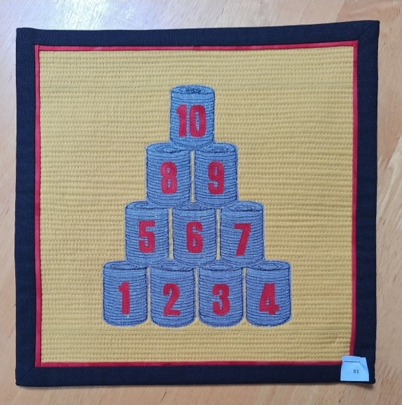 A square quilt with a yellow background and a black border. The quilt features a pyramid of gray tin cans, each with a number embroidered on it. The background is quilted with a linear pattern.