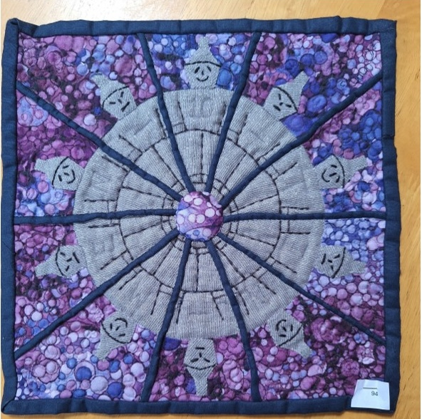 A square quilt with a purple background and a blue border. The quilt features a circular design with multiple gray figures arranged around the edge. The centre of the circle is a smaller, circular shape.