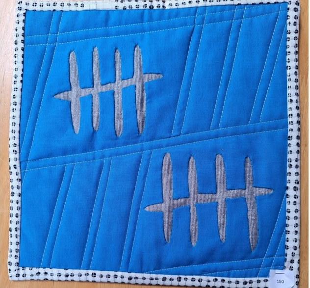 A square quilt with a blue background and a white border. The quilt features two gray, vertical lines with five horizontal lines crossing them, creating a pattern of small squares. The background is quilted with a diagonal line pattern.