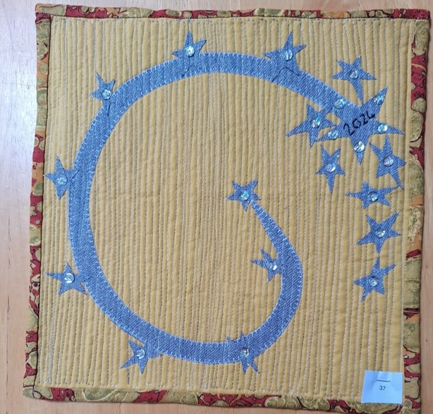 A square quilt with a yellow background and a red border. The quilt features a blue, curved line decorated with silver stars. The number "2020" is embroidered on one of the stars. The background is quilted with a linear pattern.