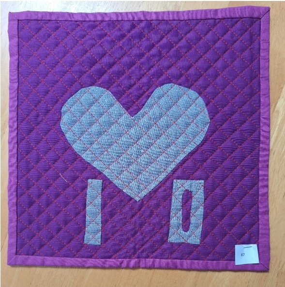 A square quilt with a purple background and a purple border. The quilt features a large, gray heart shape in the centre, with the letters "I" and "O" below it. The background is quilted with a diagonal line pattern.