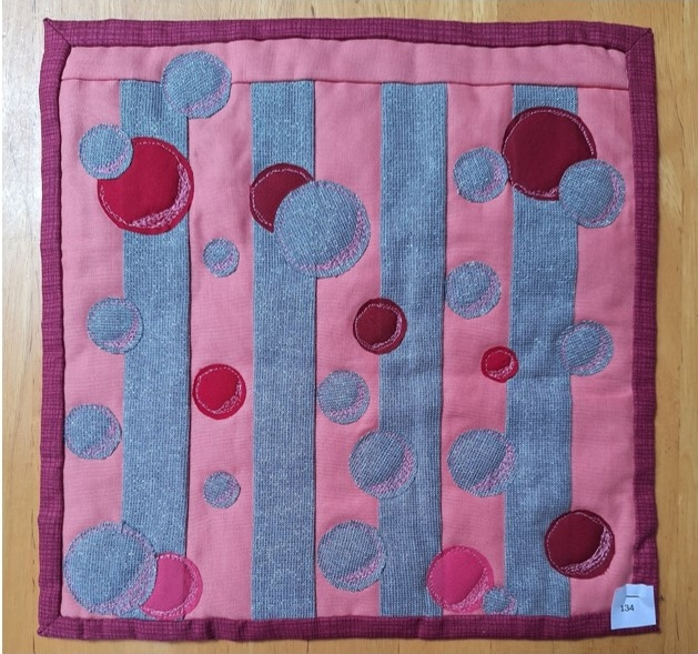 A square quilt with a pink background and a red border. The quilt features a pattern of vertical gray stripes with overlapping circles in various shades of pink and red.