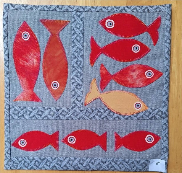 A square quilt with a gray background and a blue border. The quilt features a pattern of red and orange fish with white eyes, arranged in a grid-like pattern.