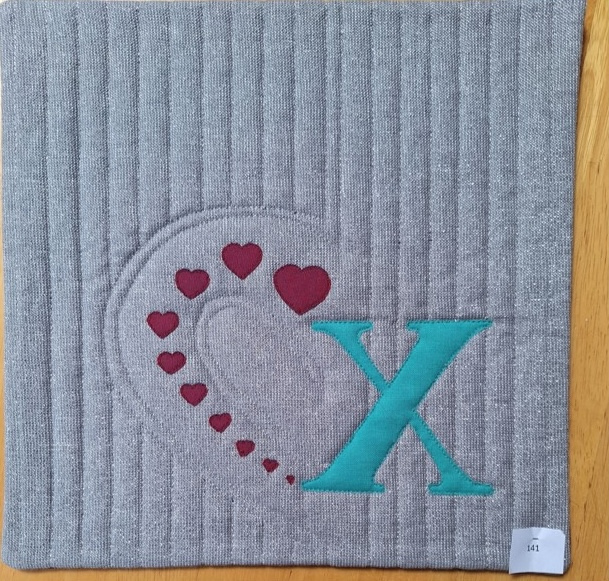 A square quilt with a gray background. The quilt features a large, blue letter "X" and a curved line of red hearts. The background is quilted with vertical lines.