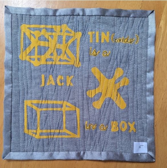 A square quilt with a gray background and a gray border. The quilt features the text "TIN (oxide) is a JACK in a BOX" in yellow letters, along with a drawing of a jack-in-the-box. The background is quilted with a linear pattern.