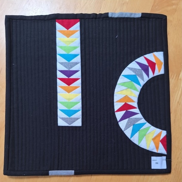 A square quilt with a black background and a black border. The quilt features a vertical and a curved arrangement of colourful triangles, creating a visually dynamic composition. The background is quilted with a linear pattern.