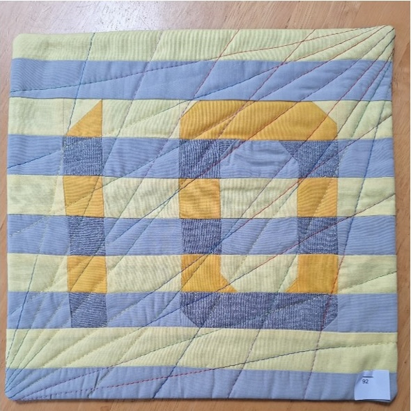 A square quilt with a yellow and blue striped background. The number "10" is formed using gray and yellow fabric pieces, with the "1" being larger than the "0". The background is quilted with a diagonal line pattern.