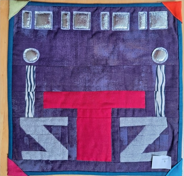 A square quilt with a purple background and a colourful border. The quilt features a stylised table and chairs, with a red table and gray chairs.