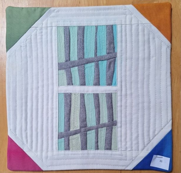 A square quilt with a white background and a colourful border. The quilt features two rectangular windows with a grid-like pattern inside. The background is quilted with a linear pattern.