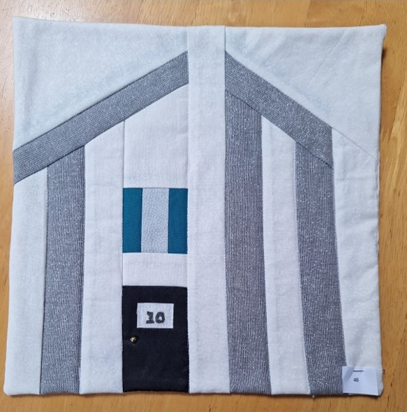 A square quilt with a white background and a gray border. The quilt features two houses, each with a door and a window. The houses are made from gray fabric with blue and green details for the door and window.