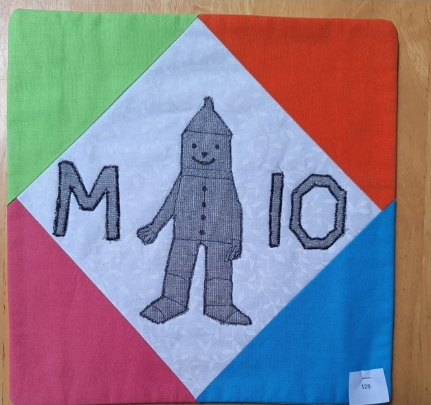 A square quilt with a colourful background and a white centre. The centre features a gray, embroidered figure of the Tin Man from The Wizard of Oz, along with the letters "M" and "10". The background is divided into four triangular sections in green, red, blue, and pink