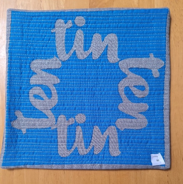 A square quilt with a blue background and a gray border. The word "TIN" is repeated four times in a circular pattern, with each letter made of gray fabric. The background is quilted with a linear pattern.