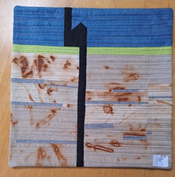 A square quilt with a blue background and a black border. The quilt features a vertical black line dividing the background into two sections. The top section is blue with a yellow line, while the bottom section is a patchwork of white and brown fabric with a rusty appearance. The background is quilted with a linear pattern.