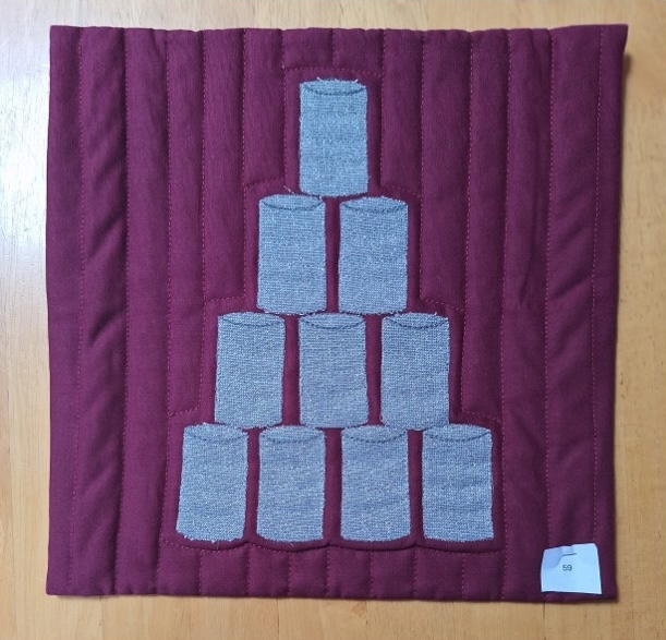 A square quilt with a red background and a red border. The quilt features a pyramid of gray tin cans, arranged in a triangular pattern. The background is quilted with vertical lines.