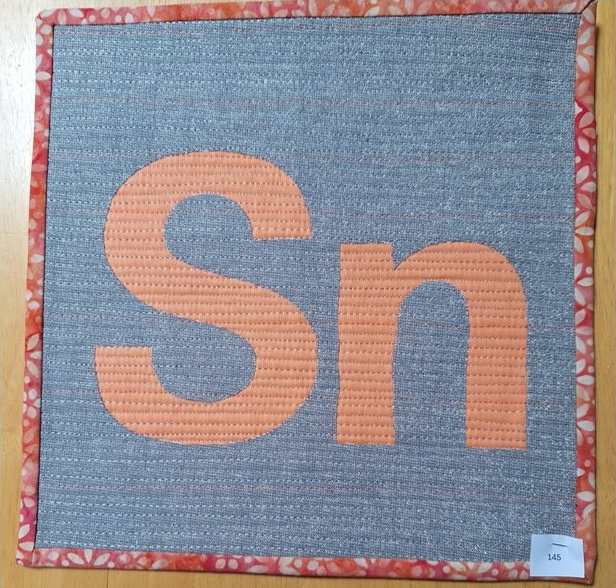 A square quilt with a gray background and an orange border. The quilt features the letters "Sn" in orange, with a textured background. The background is quilted with matchstick lines.