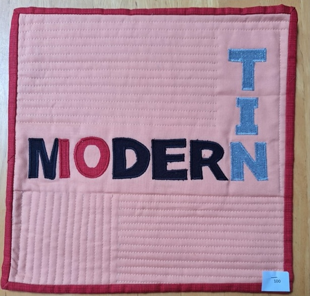A square quilt with a pink background and a red border. The quilt features the word "MODERN" spelled out in colourful letters, with the word "TIN" vertically stacked on the right side. The background is quilted with a linear pattern.